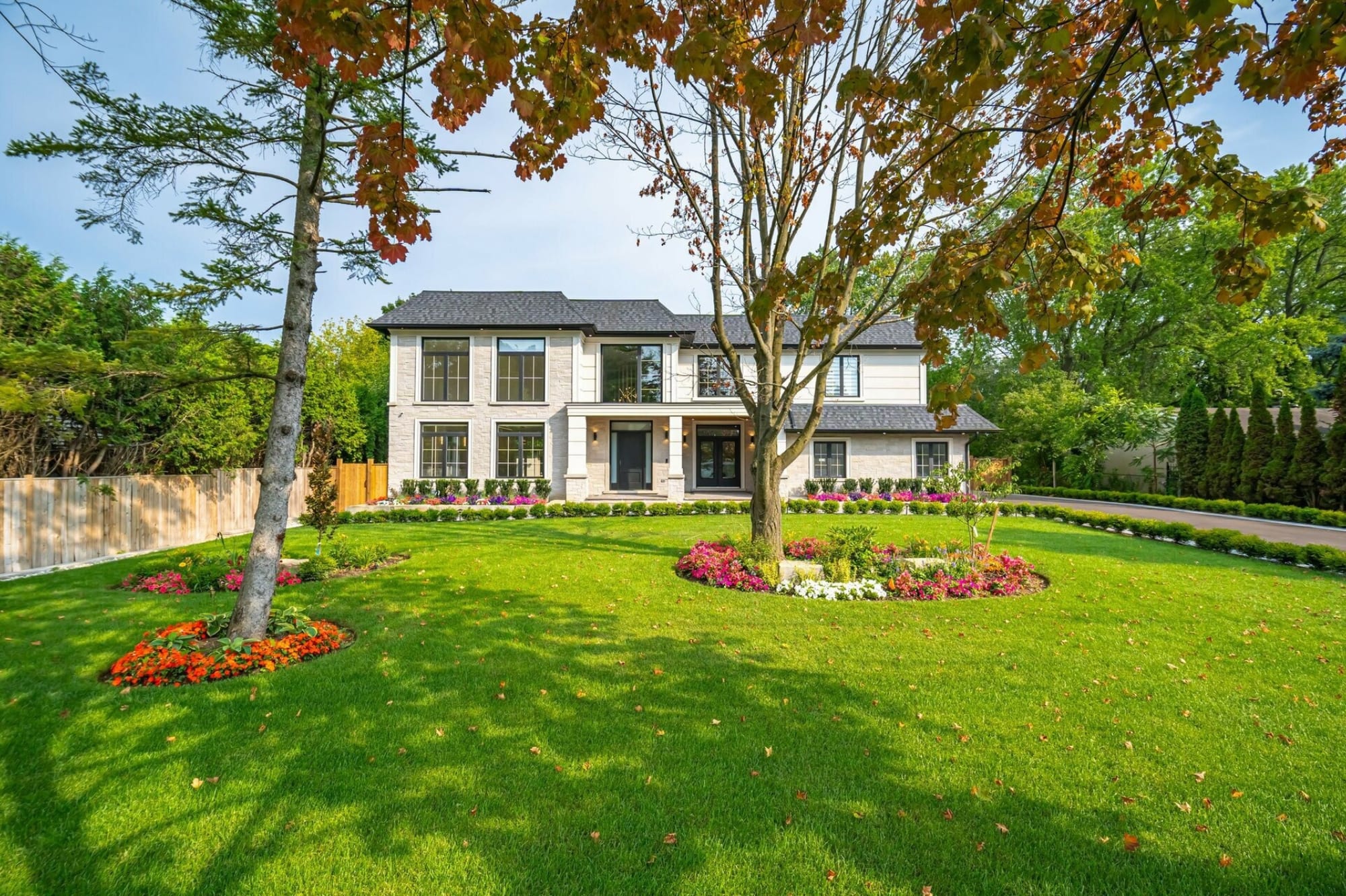 Contemporary limestone-clad two-storey home with black-framed windows behind a wide lawn, flower beds and mature trees in early autumn.