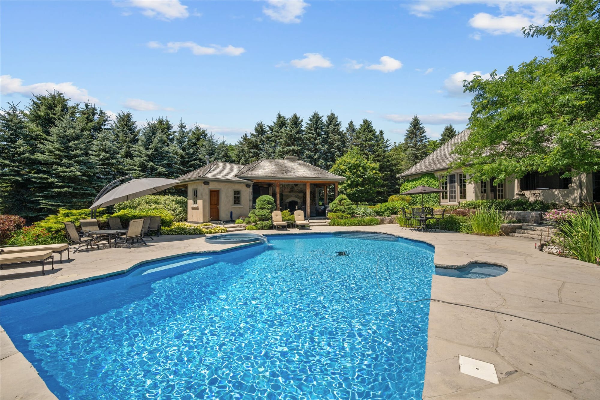 Pool and stone terrace in front of a stucco pool house with a covered loggia, framed by tall evergreens beside the main home.