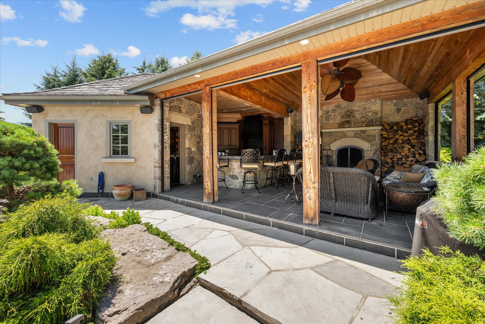 Covered outdoor living room of a pool house with a stone fireplace, a bar, wicker seating and stacked firewood.