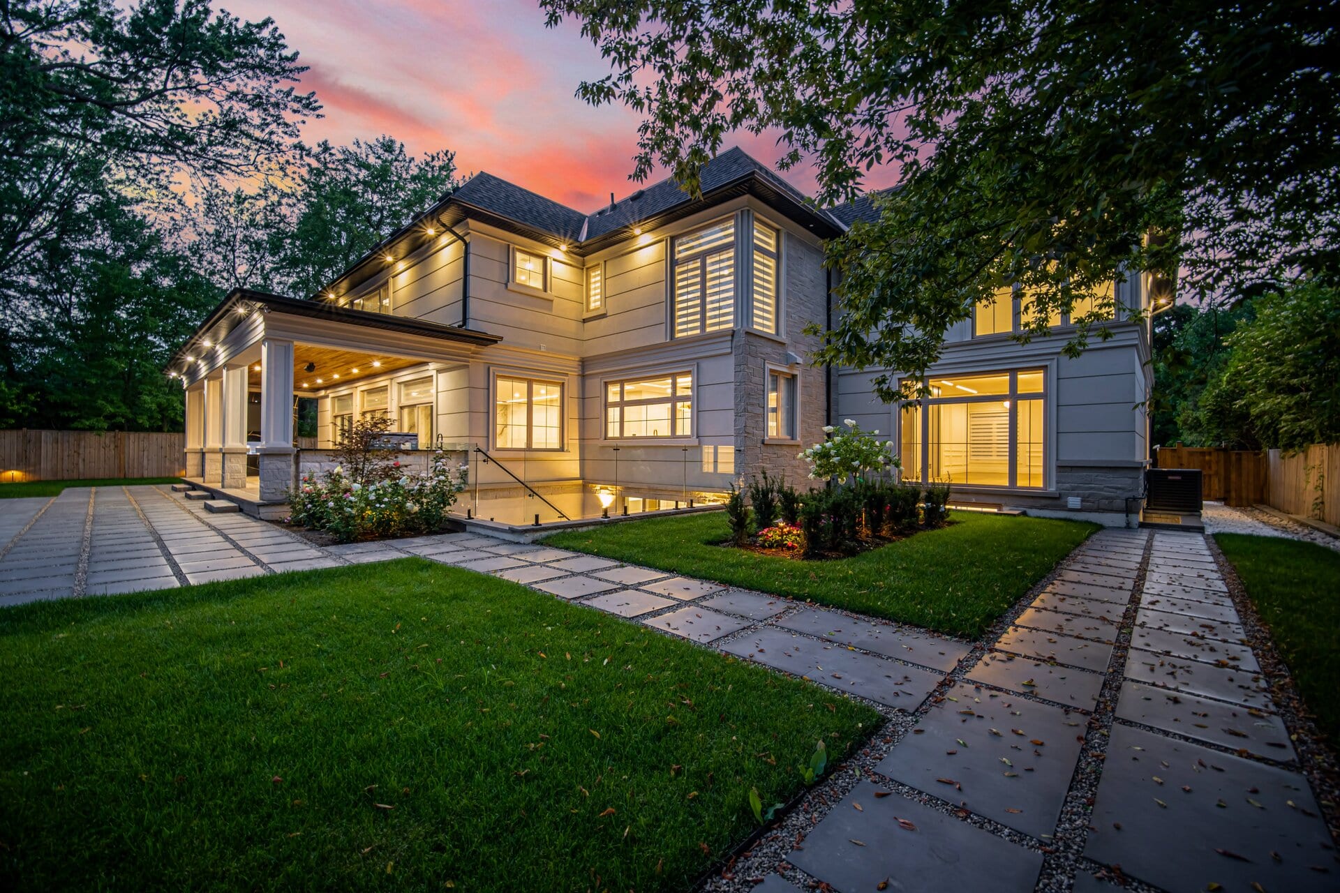 Rear of a new stone-and-siding home at dusk, with a covered loggia, a glass-railed lower terrace and a stone path across the lawn.