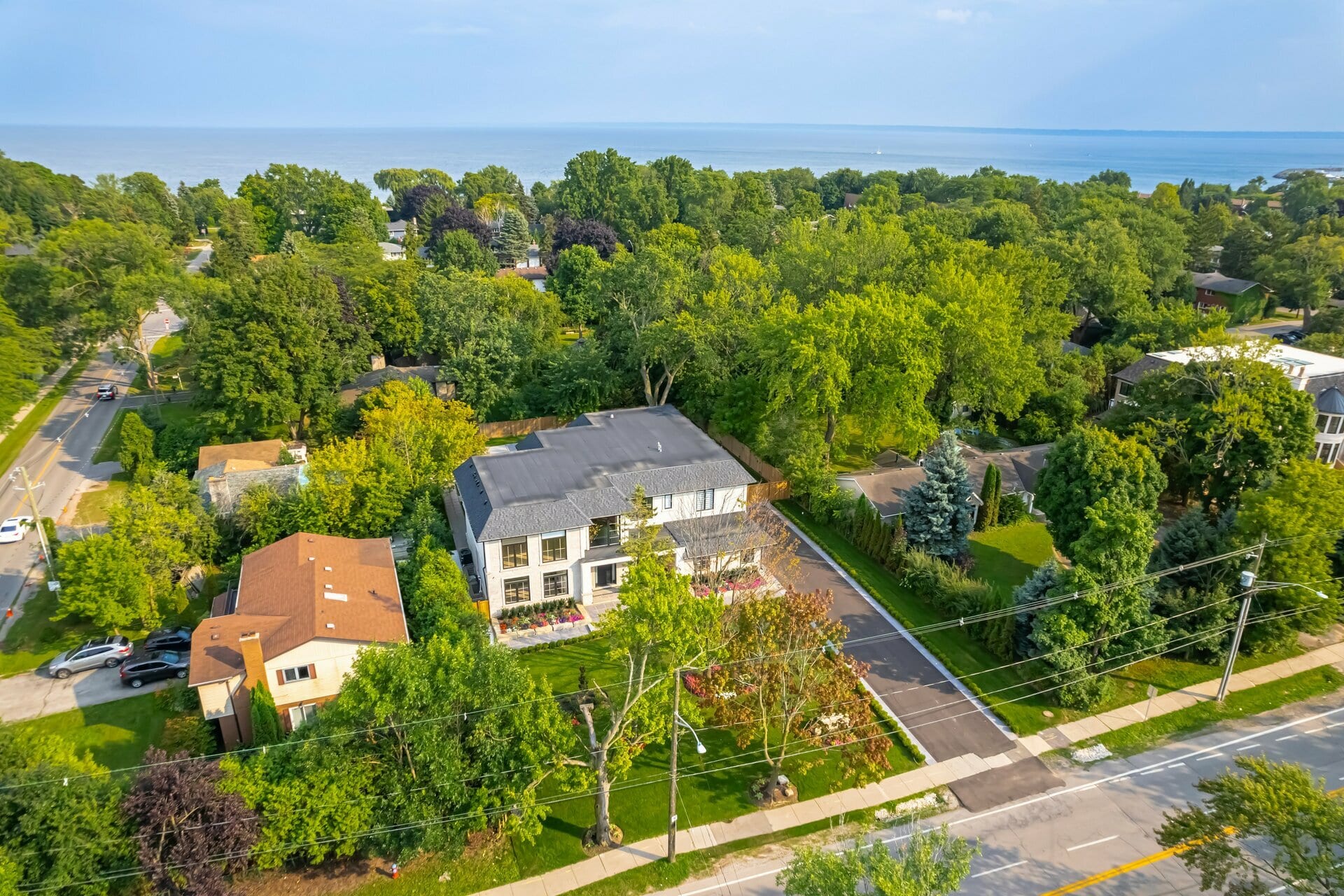 Aerial view of a new white two-storey home on a large lot on a tree-lined street, with Lake Ontario in the distance.