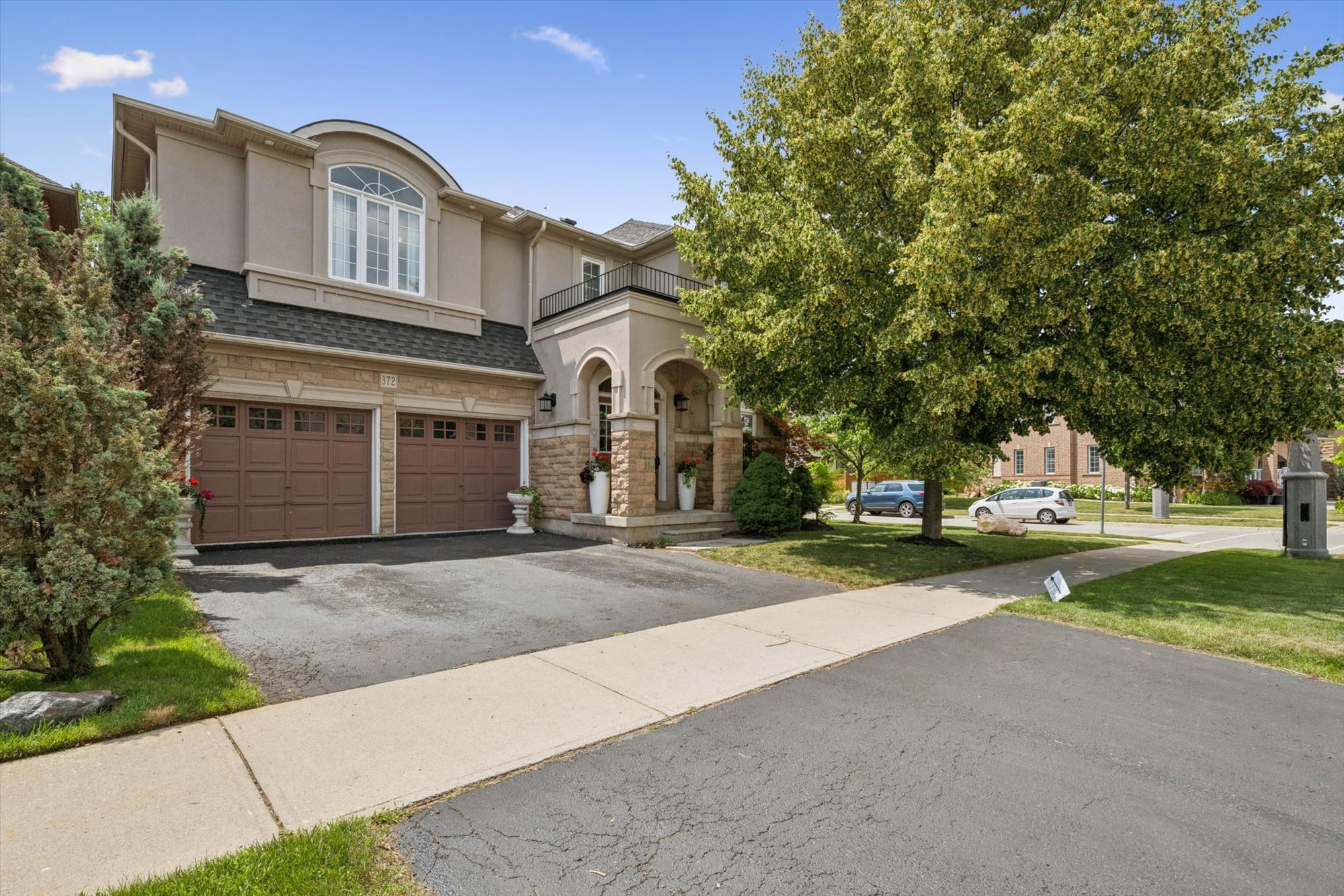 Stucco and stone two-storey home with a double garage and an arched front porch beside a mature street tree.