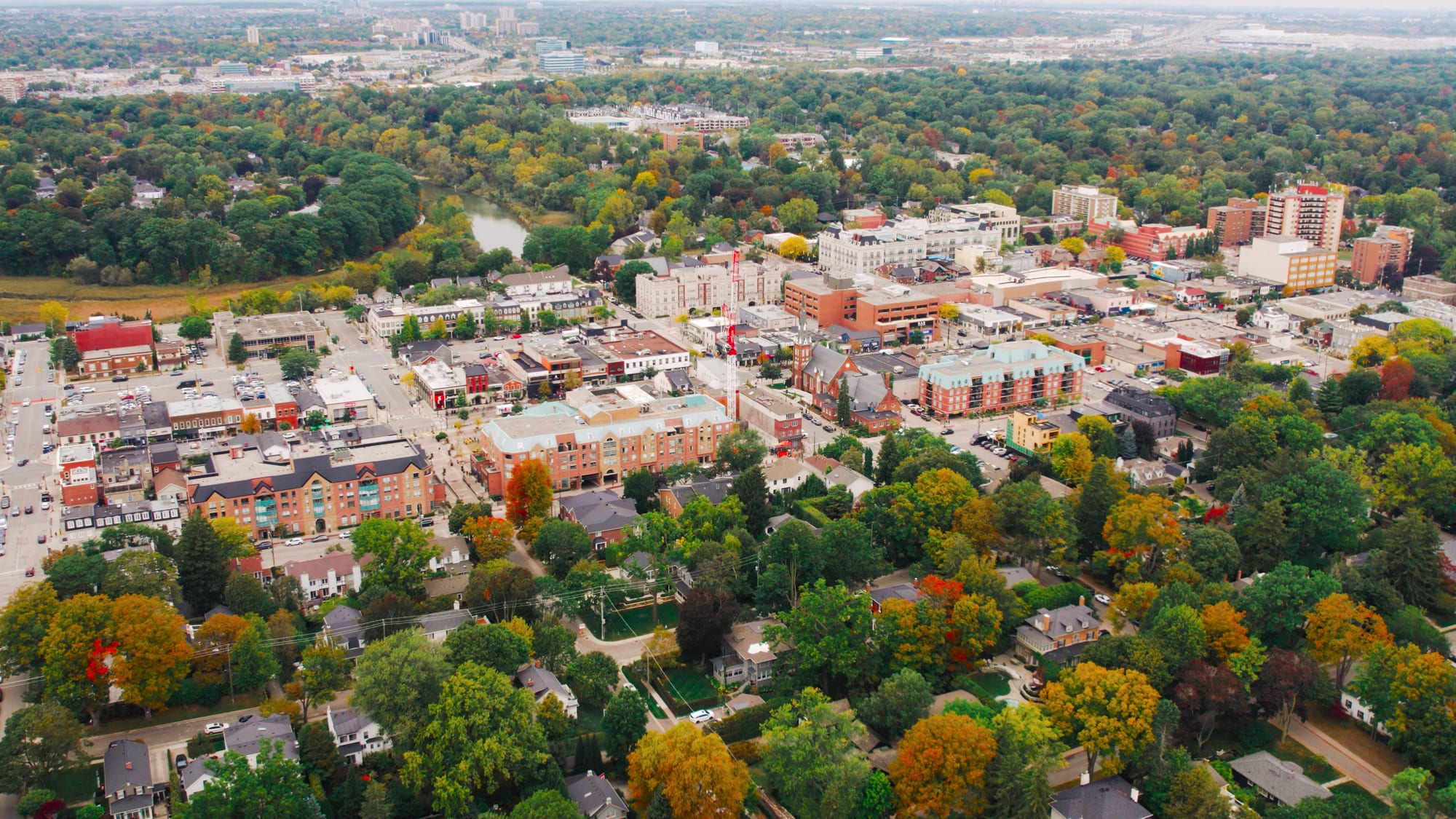 Aerial view of a downtown of low-rise shops and condominiums beside a creek, framed by autumn trees.
