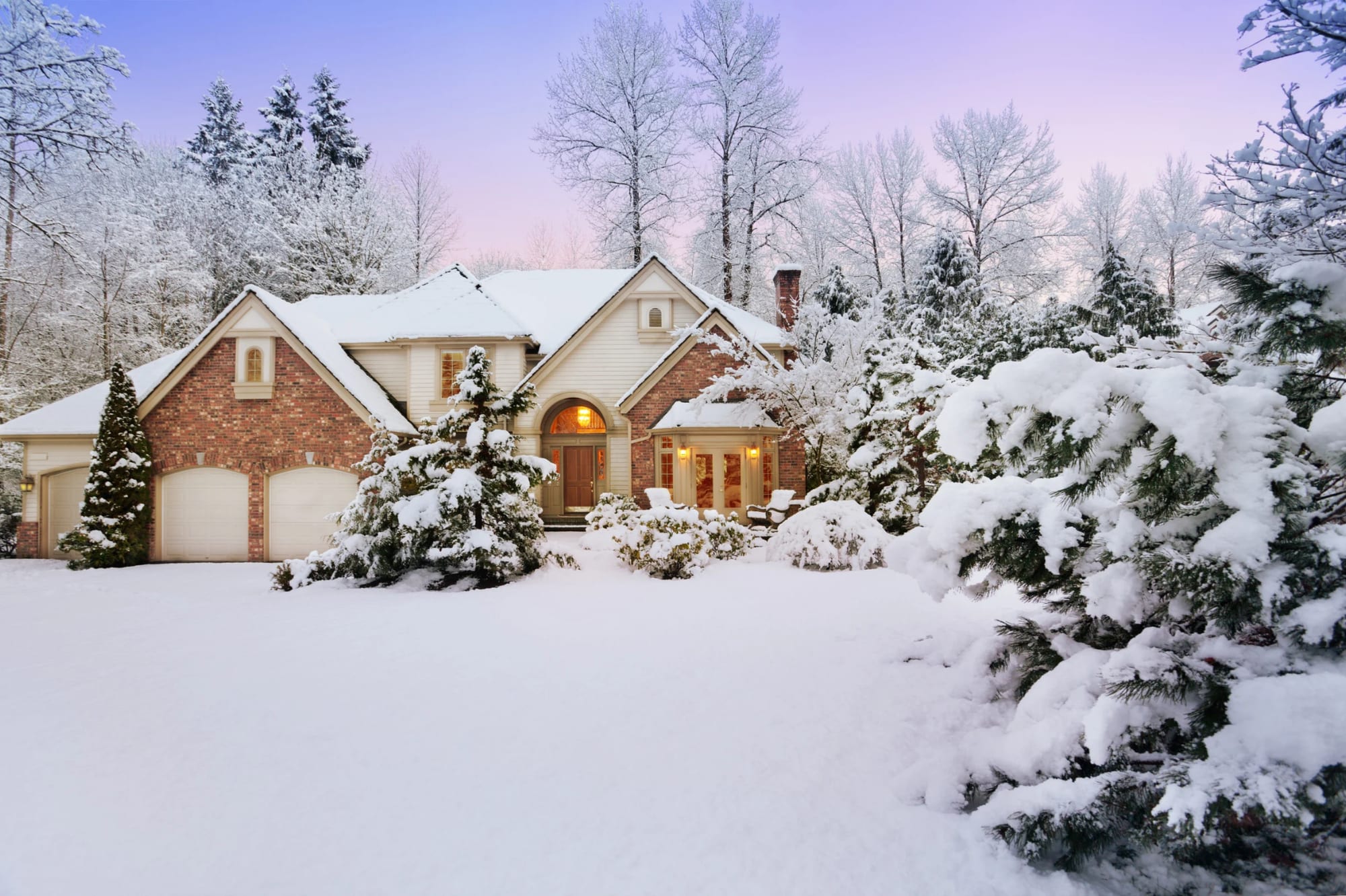Brick home with a three-car garage under fresh snow at dusk, framed by snow-covered evergreens.