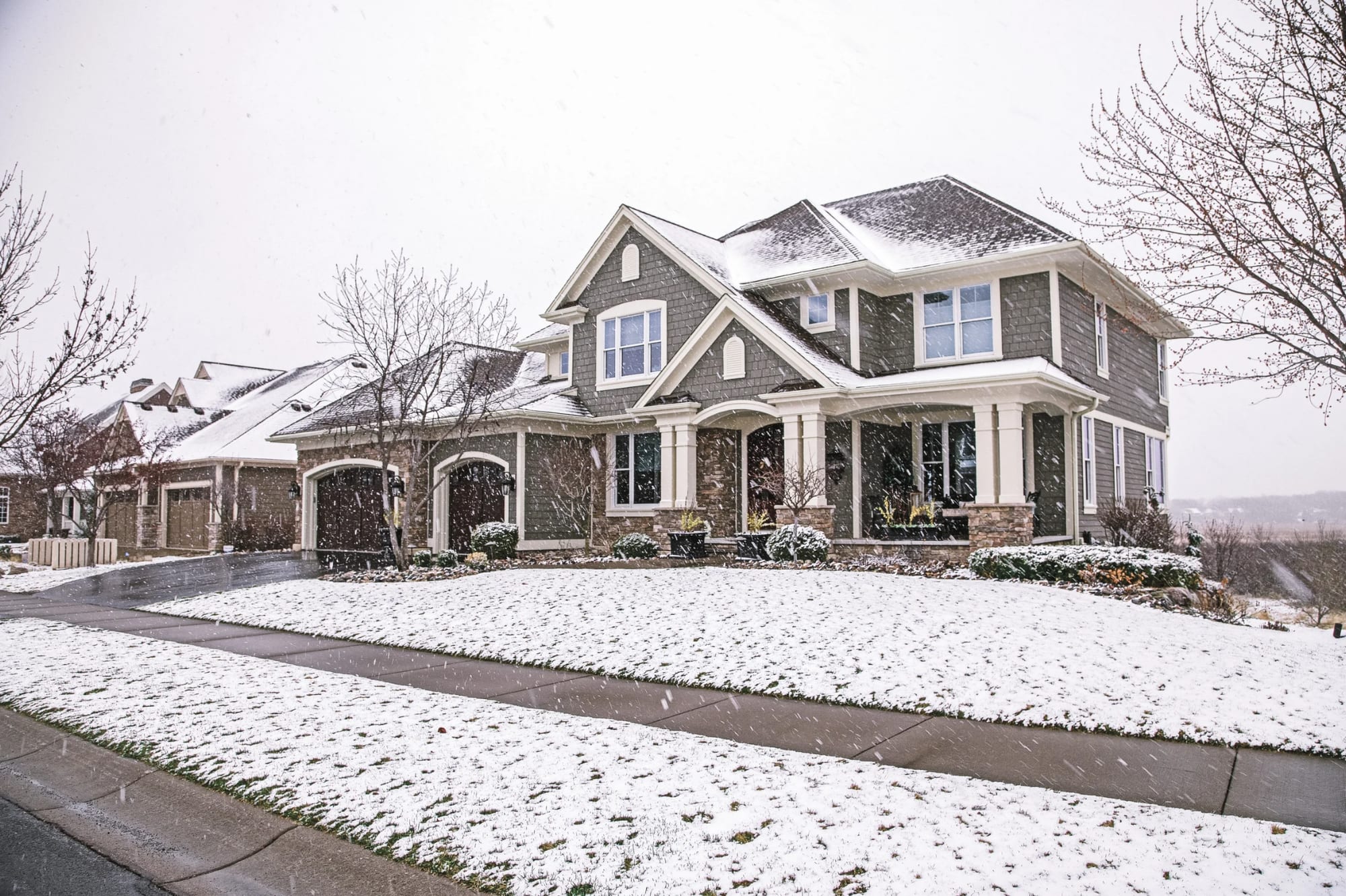 Craftsman-style home with shingle siding and stone-based columns as light snow falls on the front lawn.