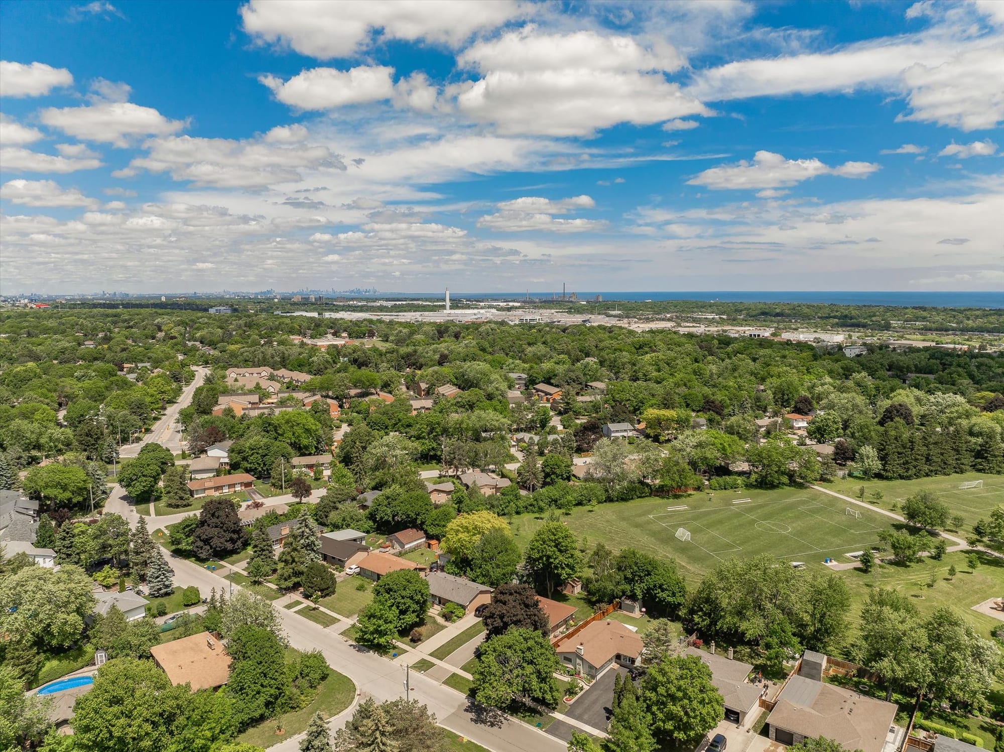 Aerial view over a leafy residential neighbourhood and sports fields, with Lake Ontario and the Toronto skyline on the horizon.