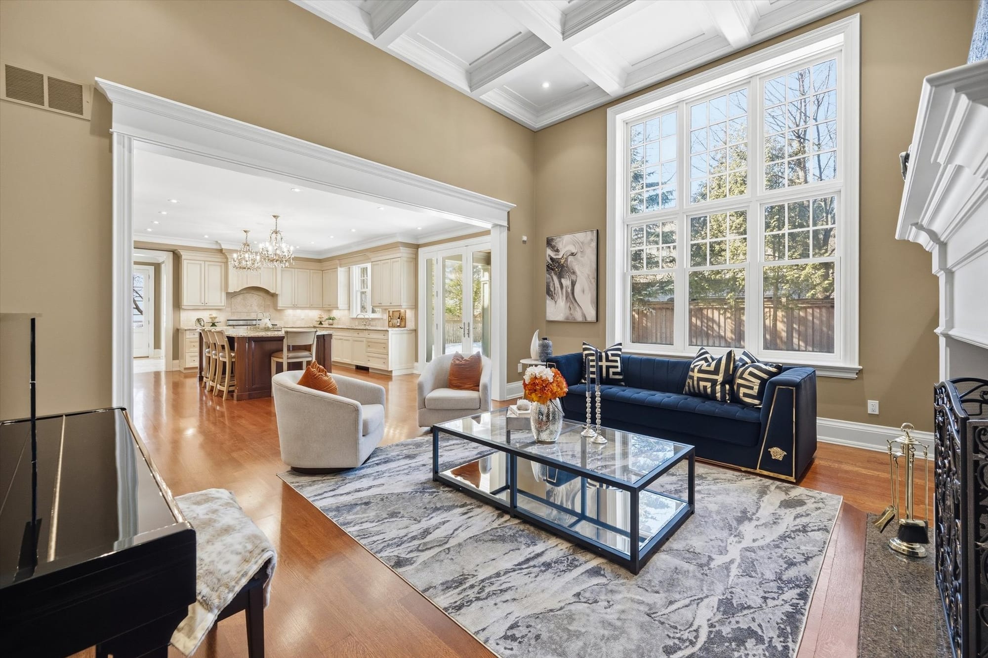 Elegant living room with a coffered ceiling and tall windows, opening onto a traditional kitchen.