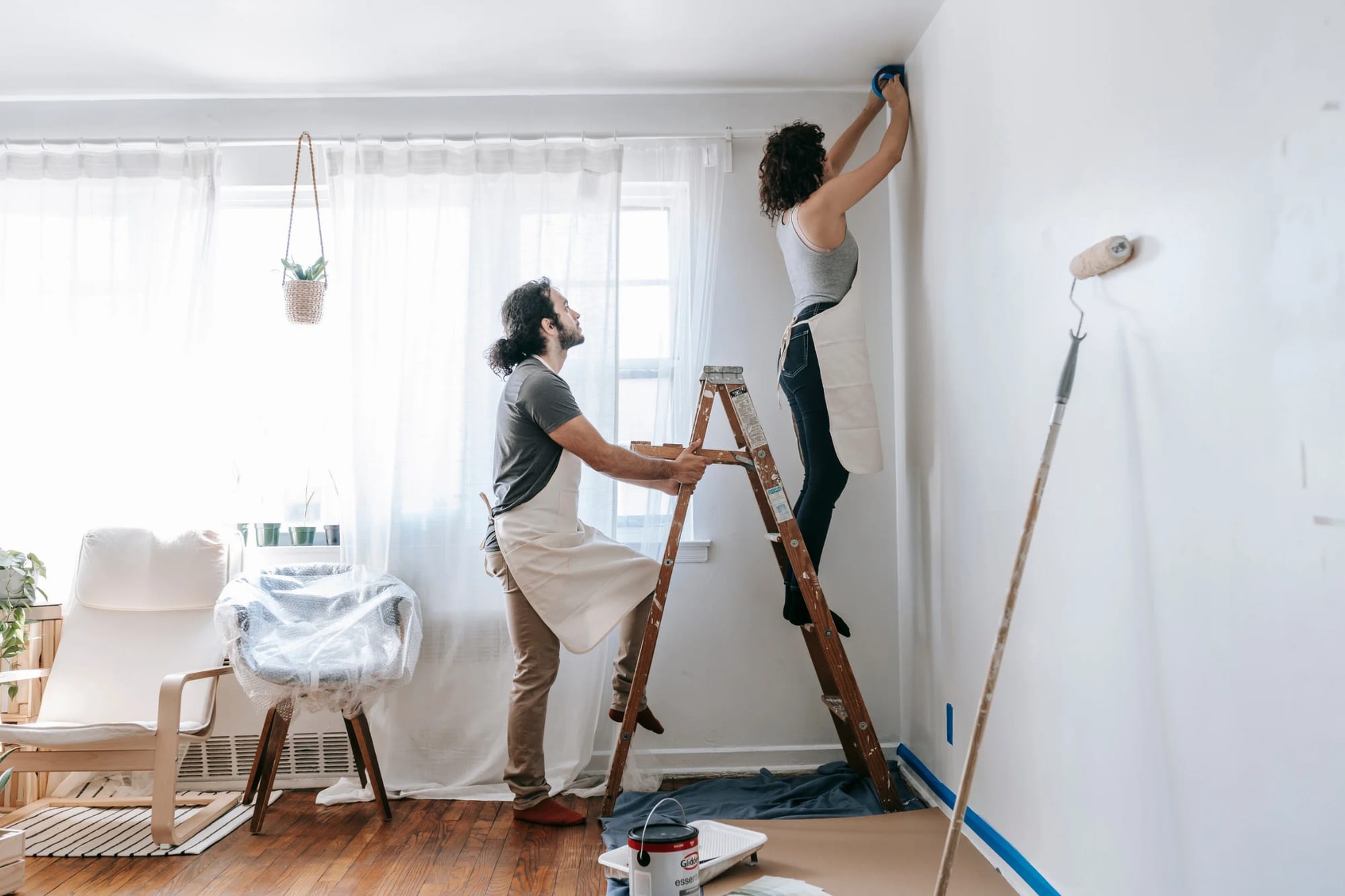 Couple painting a bright living room, one on a stepladder taping the top of a wall.